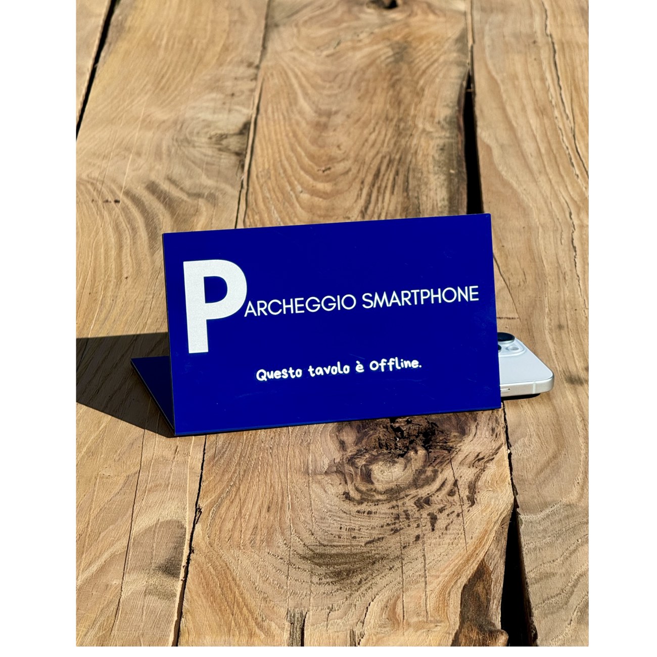 Il segnale TTIO «Parcheggio Smartphone — Questo tavolo è Offline» su un tavolo di legno, con uno smartphone posato accanto.