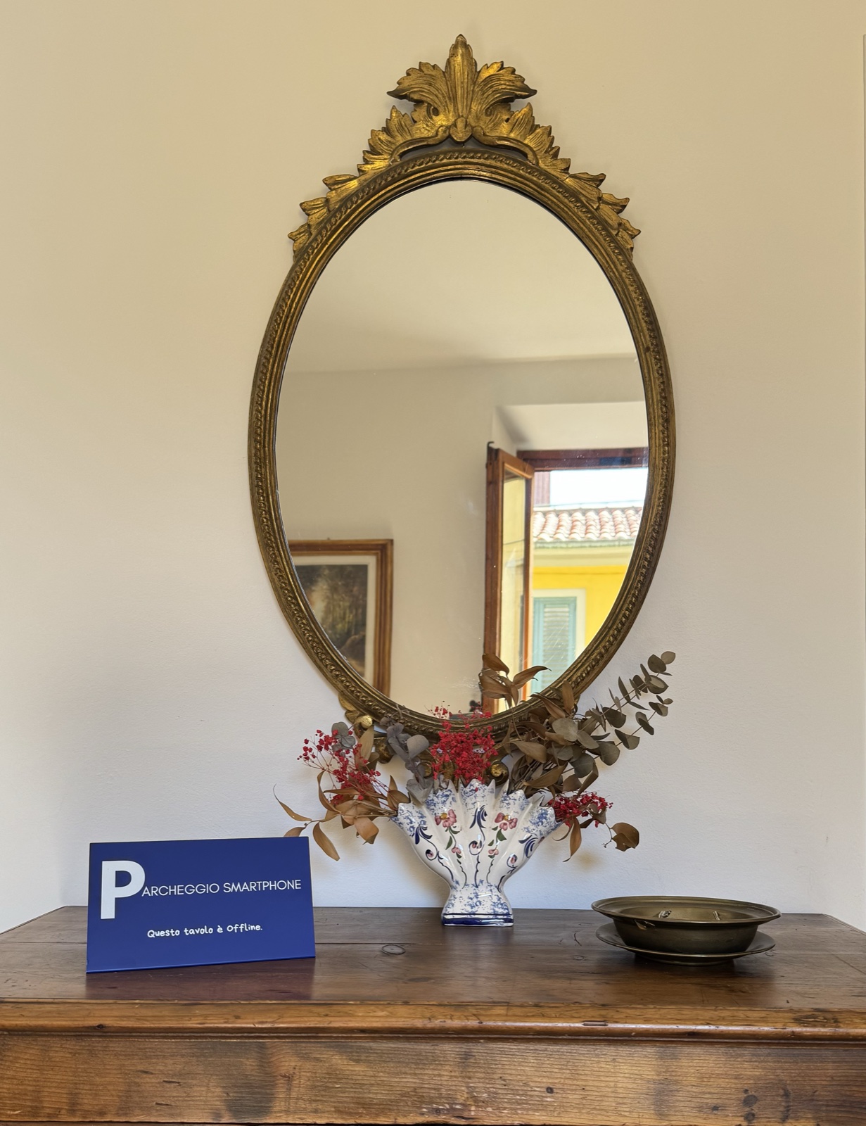Il segnale TTIO Home «Parcheggio Smartphone — Questo tavolo è Offline» su una consolle di legno, sotto uno specchio ovale dorato con un vaso di fiori.